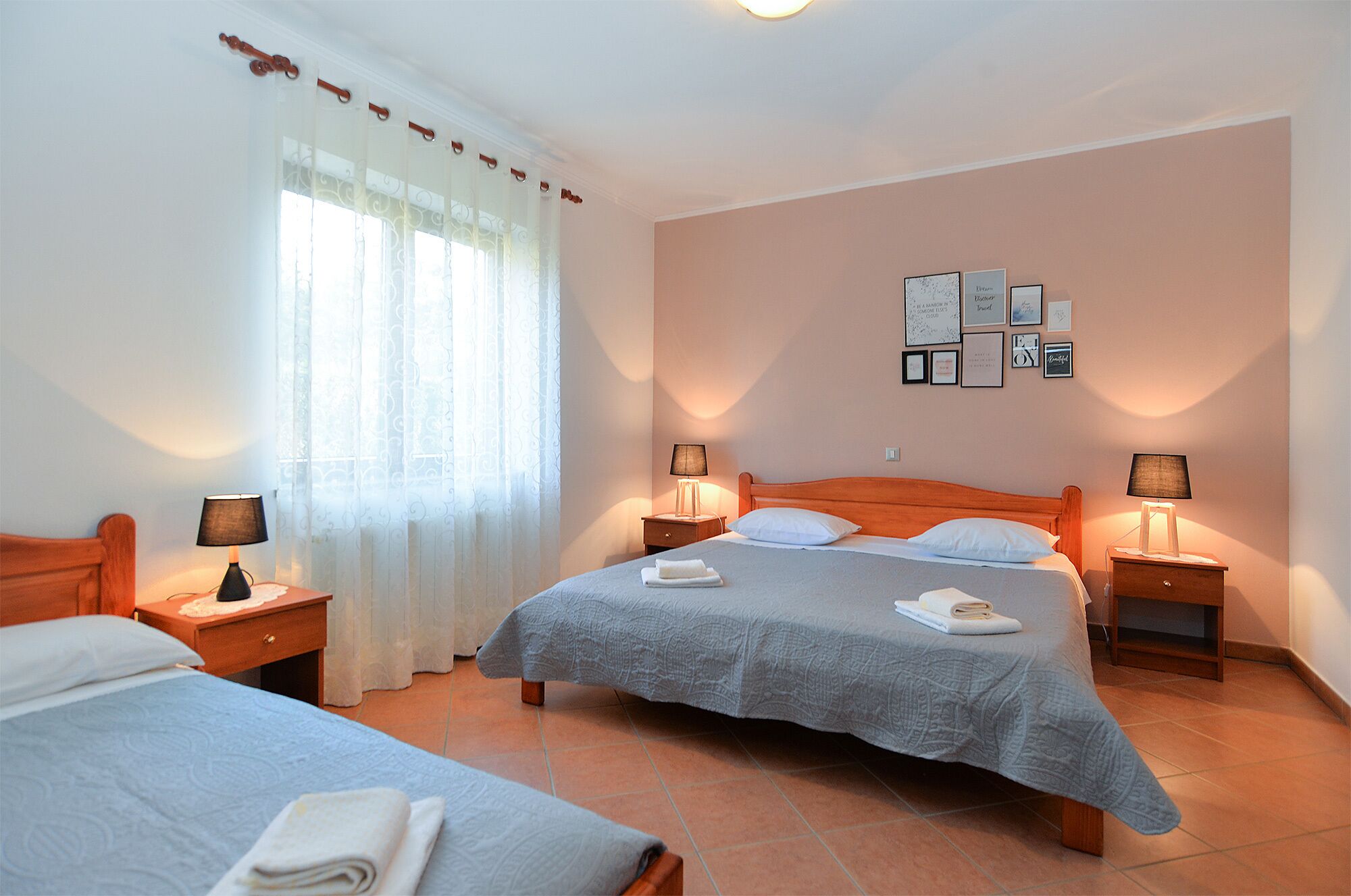 A hotel room with two beds, a large window with curtains, bedside lamps, and a wall with framed pictures.