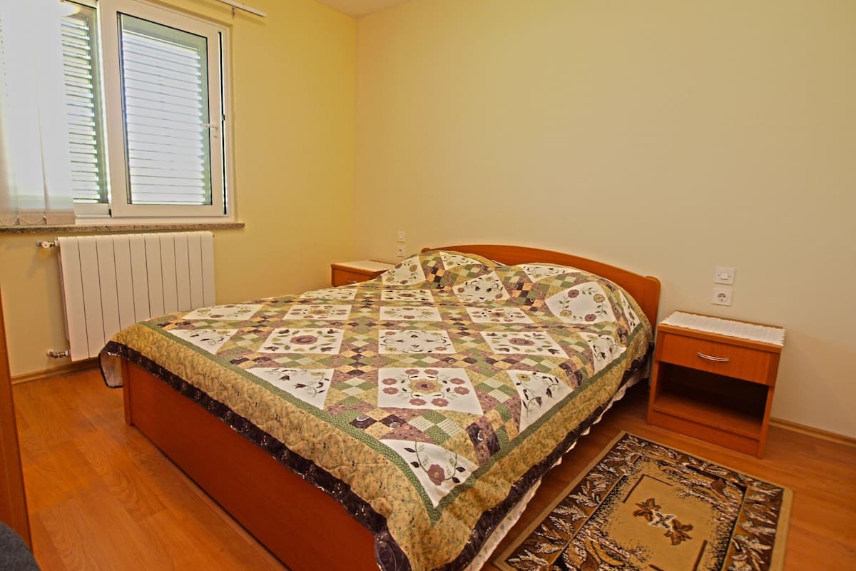 A bedroom with a wooden bed, a nightstand, a bedside table, a window with blinds, and a radiator.