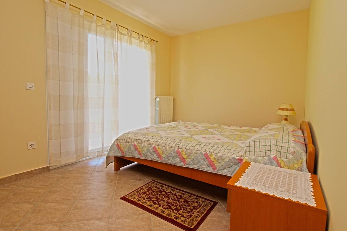 A bedroom with a bed, a nightstand, a lamp, a window with curtains, and a radiator.