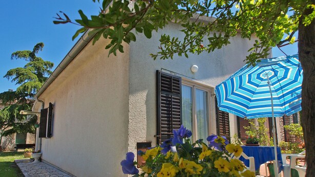 A house with a stone pathway leading to a door, a blue-striped umbrella, and a garden with yellow and blue flowers. - Apartment 1095 (Pula)