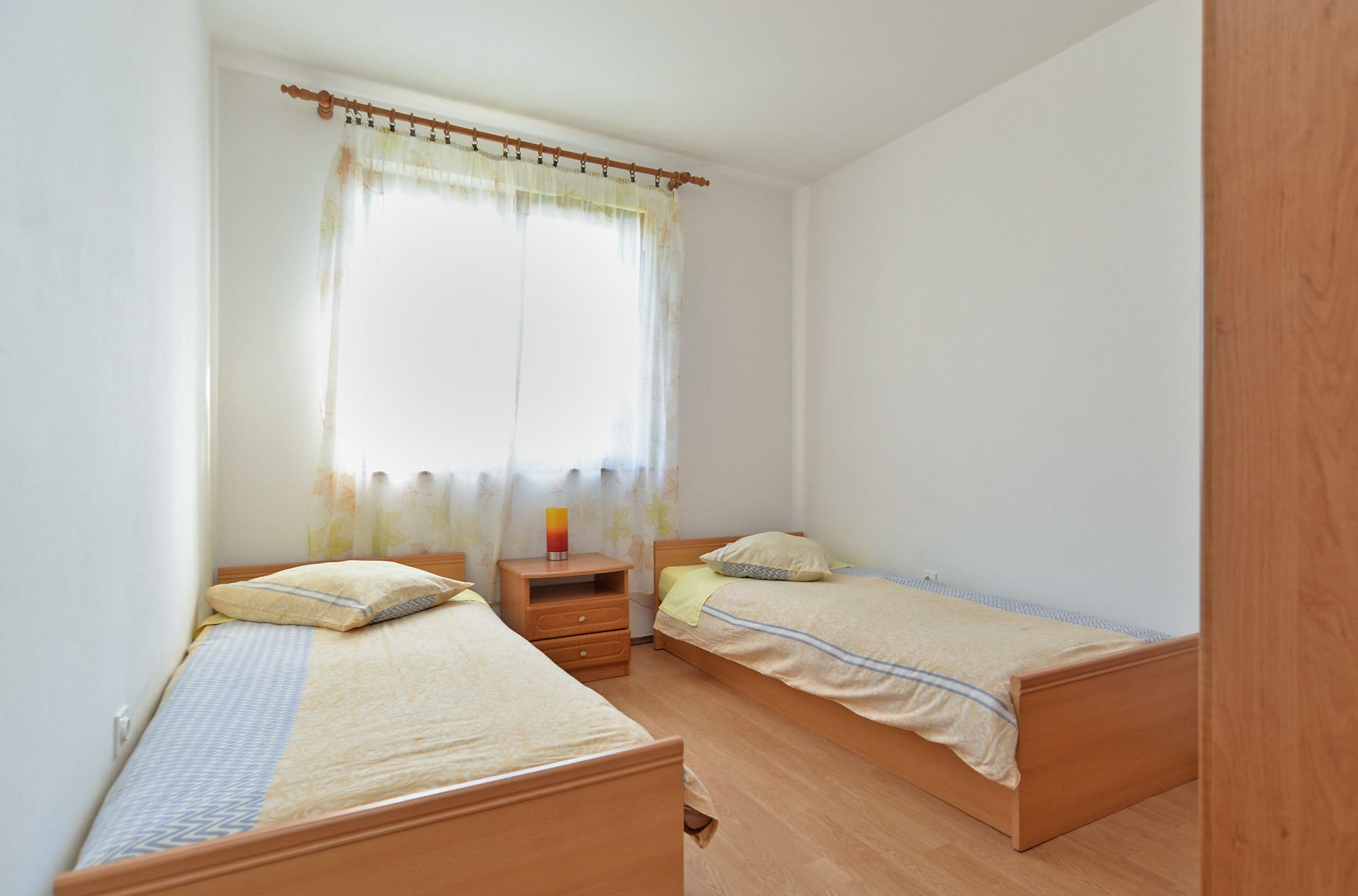 A small, clean room with two wooden beds, a nightstand, and a window with a curtain.