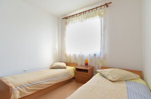 A bedroom with two single beds, a wooden nightstand, a bedside table, and a window with sheer curtains.