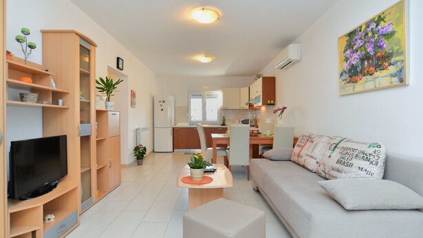A modern living room with a flat-screen TV, wooden shelving, a sofa, and a dining area with a kitchen in the background. - Apartment 1100 (Pula)