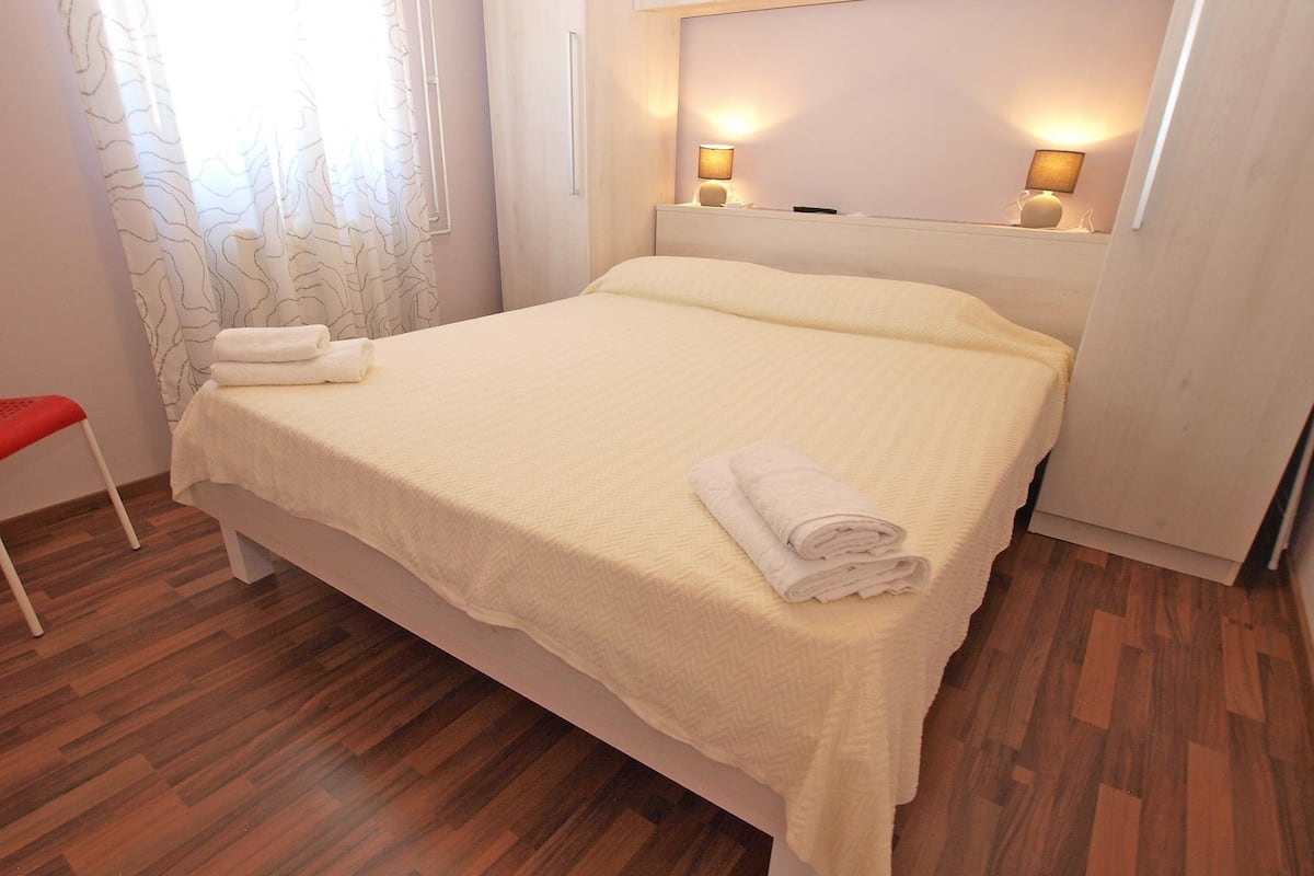 A neatly made bed with a white bedspread and two rolled towels on it. There is a wooden floor and a red chair to the left.