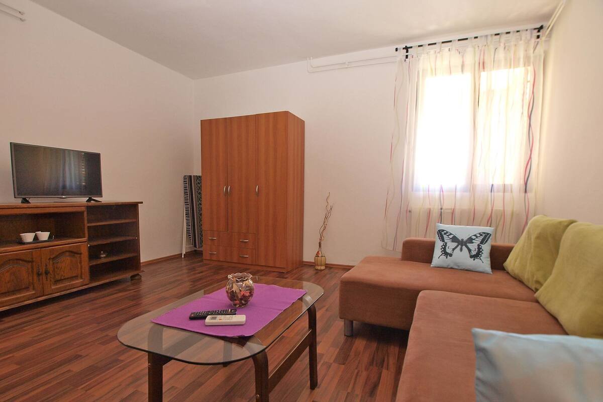 A living room with a sofa, a glass coffee table, a TV on a wooden stand, a wardrobe, and a window with curtains.