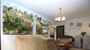 A living room with a beige sofa, a patterned armchair, and a floral sofa. There are several potted plants on a shelf and hanging from the ceiling. A chandelier hangs from the ceiling, and a clock is visible on the wall. - Apartment 1123 (Vodnjan)