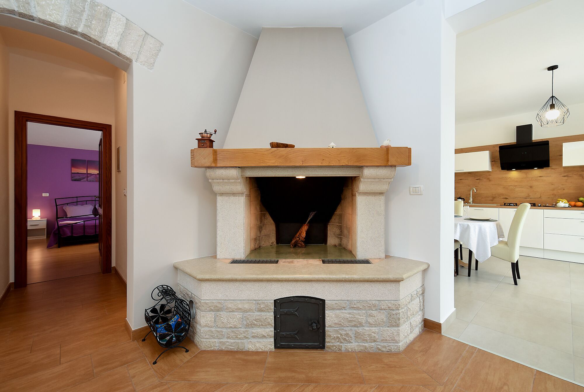 A modern living room with a stone fireplace, a dining area with a table and chairs, and a kitchen with a wooden countertop and a hanging light fixture.