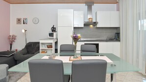 Uma sala de estar moderna com uma mesa de jantar de vidro, sofás cinzas e uma cozinha com armários brancos e painel de fundo cinza.