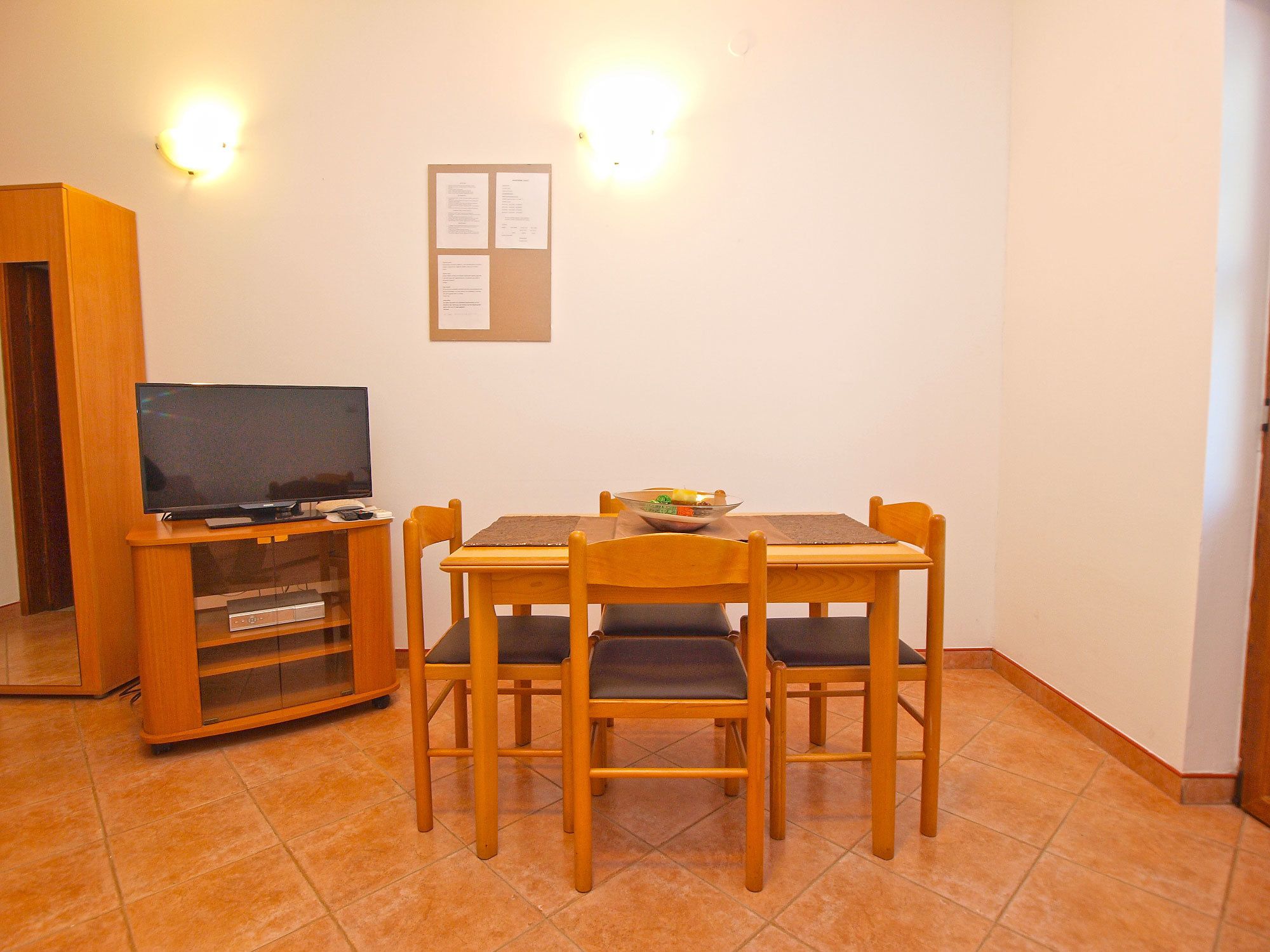 Een eetgedeelte met een houten tafel, vier stoelen, een televisie op een standaard en een prikbord aan de muur.