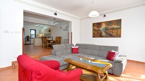 A living room with a grey sofa, red armchairs, and a wooden coffee table. There is a painting on the wall and a kitchen in the background. - Apartment 1168 (Fazana)