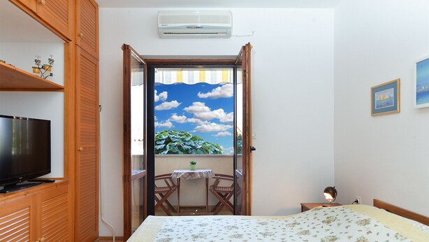 Un dormitorio con cama, televisor, una mesita con una planta y balcón con vistas a vegetación y cielo azul.