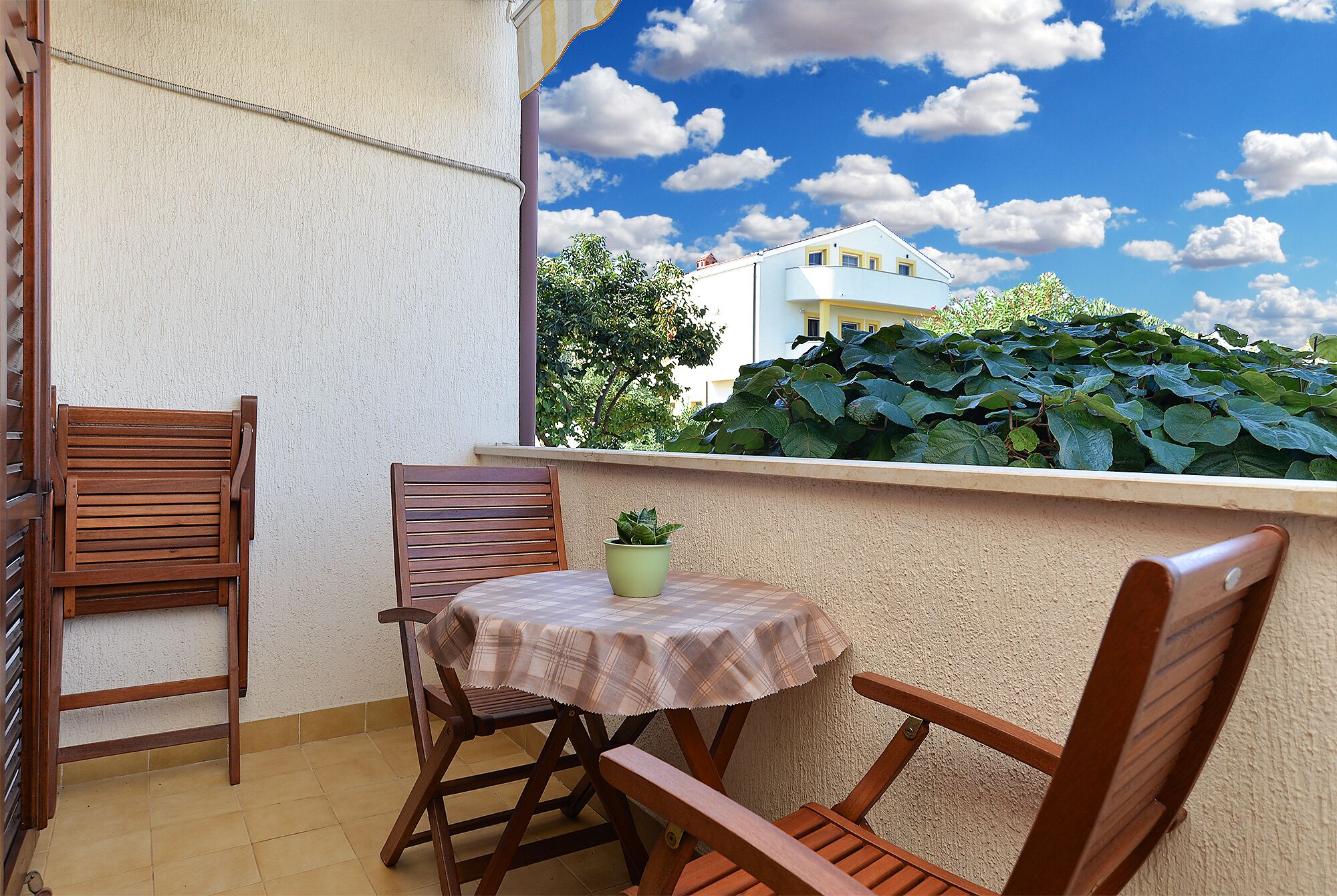 En lille balkon med et bord og stole, en potteplante og udsigt til en bygning og grøn vegetation.