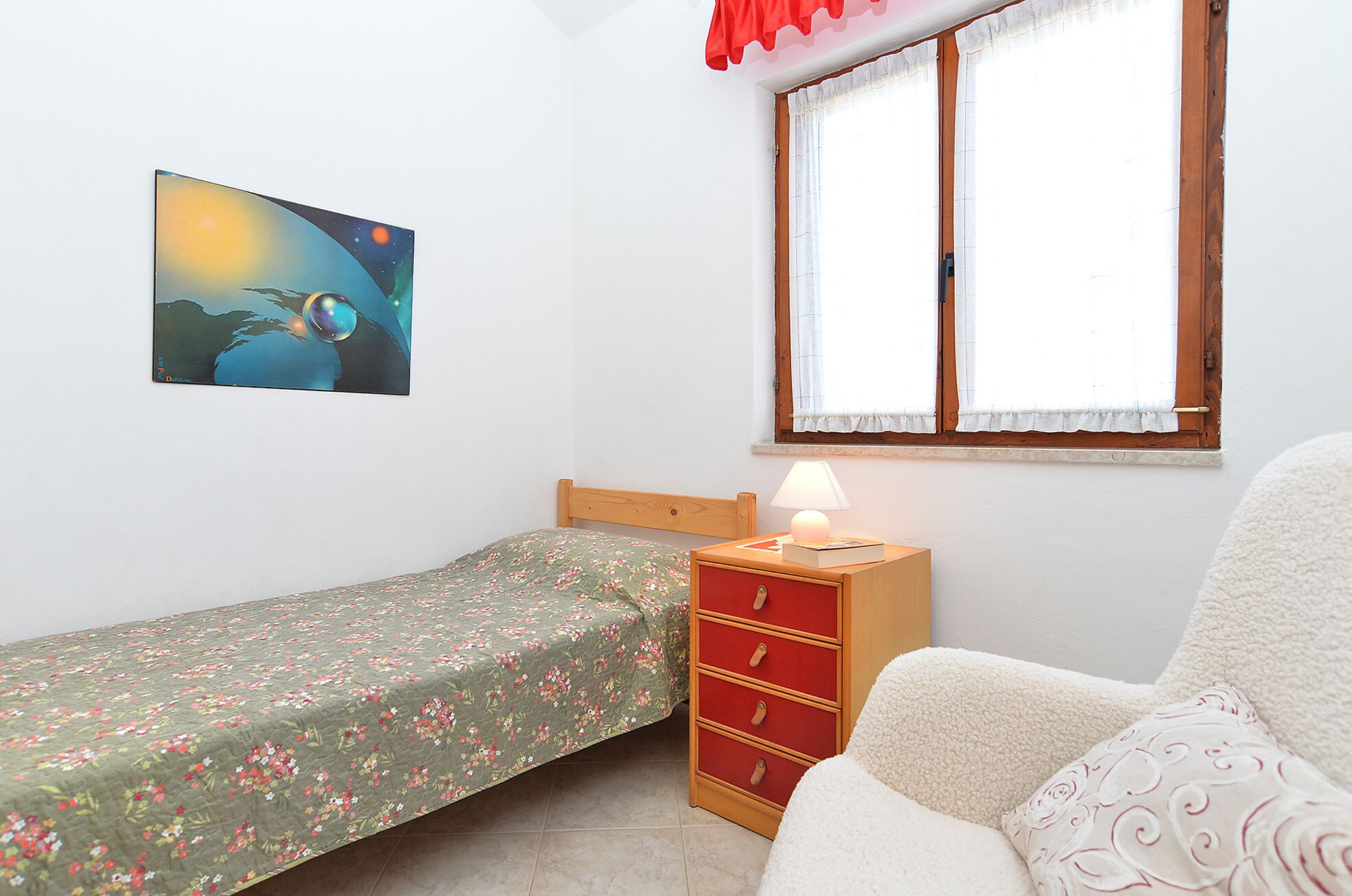 Une petite chambre à coucher, bien rangée, avec un lit simple, une table de chevet avec une lampe, une commode et une fenêtre avec des rideaux.