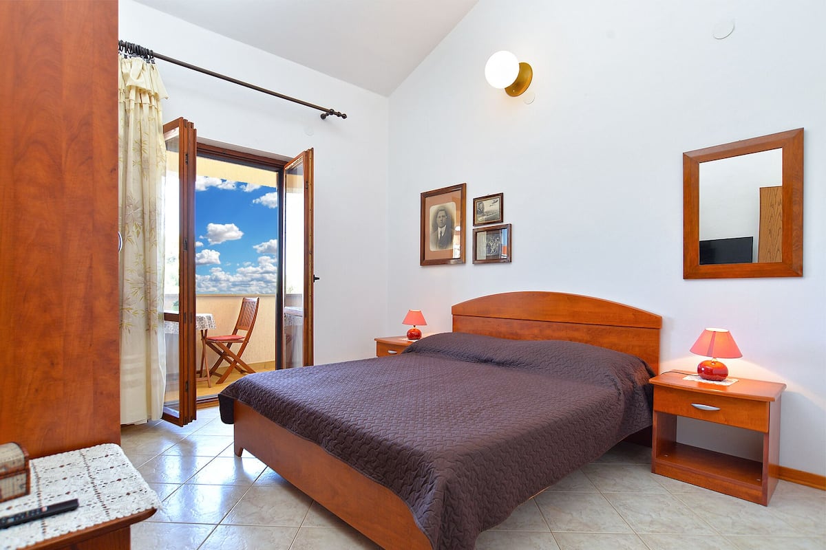 Une chambre à coucher comprenant un lit, deux tables de chevet, une chaise, une fenêtre avec des rideaux et un mur orné de tableaux.