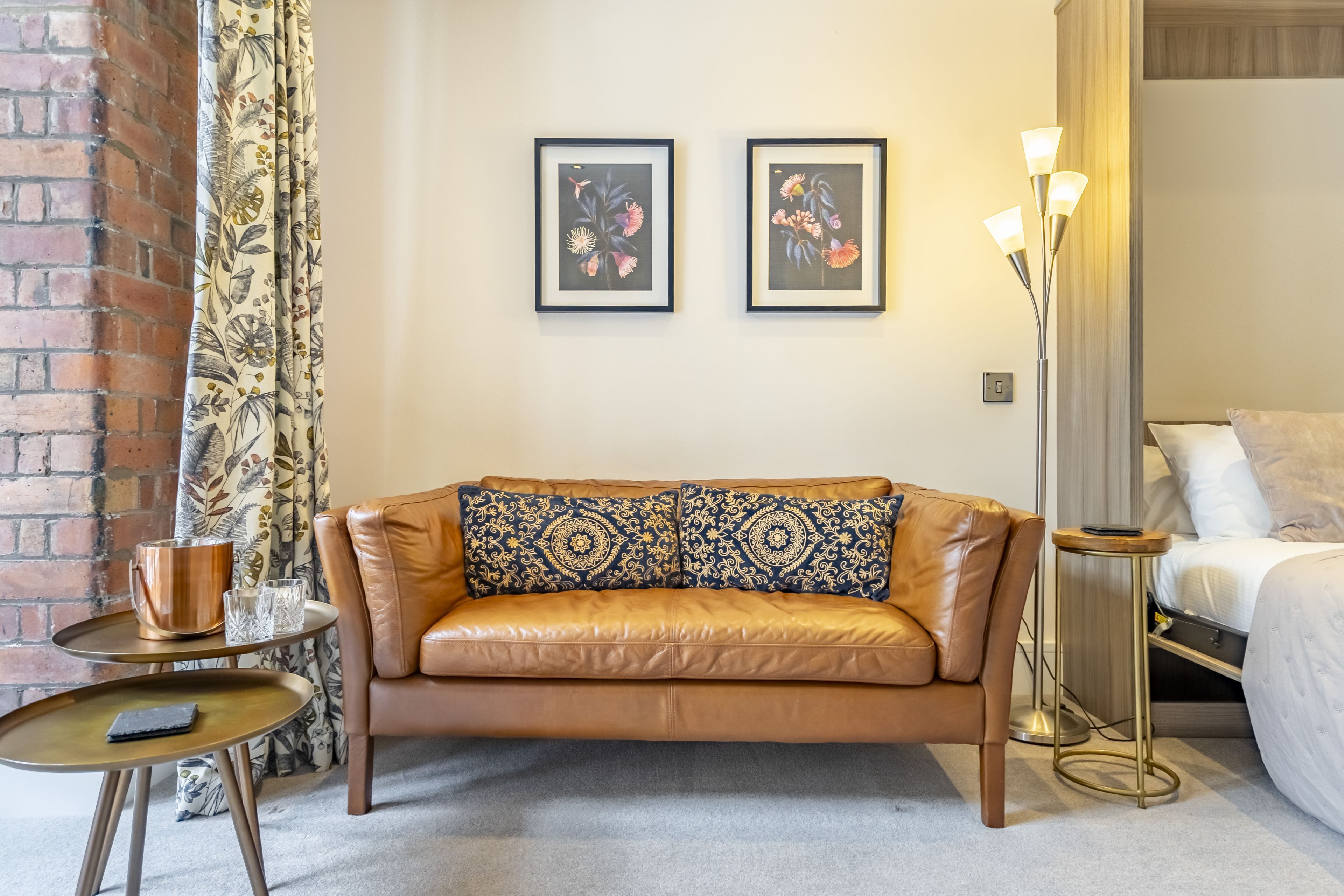 A room with a leather sofa, a small round table with a book, a floor lamp, and a bed.