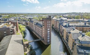 Ein Fluss fließt durch eine Stadt, gesäumt von Gebäuden, eine Brücke überspannt ihn, und in der Ferne ist die Landschaft zu sehen.