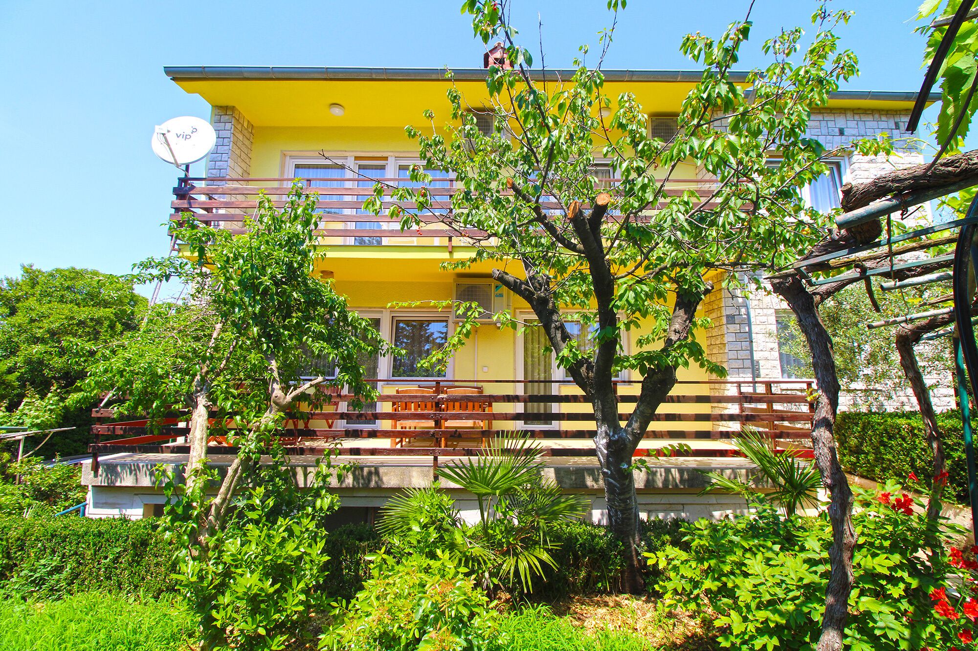 A yellow two-story building with a balcony, surrounded by lush greenery and a wooden fence.