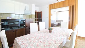 A dining area with a table set for four, surrounded by white chairs. There is a buffet counter with stacked plates and a glass display case. The room has wooden paneling on the walls and a large window with curtains. - Apartment 1198 (Medulin)