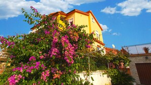 Ein gelbes Gebäude mit rotem Dach, umgeben von üppigem Grün und leuchtend pinkfarbenen Blüten.
