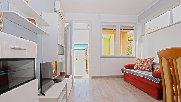 Ein modernes Wohnzimmer mit Fernseher, Sofa und Holzstuhl. Es gibt eine Küchenzeile mit Kühlschrank und Fenster.