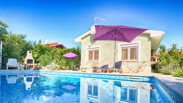 Une piscine avec des chaises longues et des parasols, un barbecue et une maison dotée d’un grand parasol.