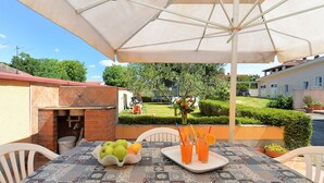 An outdoor dining area with a table set for two, a brick oven, and a view of a residential area with a lawn and trees. - Apartment 1203 (Pula)