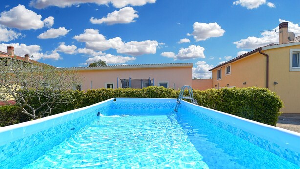 A swimming pool with clear blue water, surrounded by greenery and buildings. - Apartment 1203 (Pula)