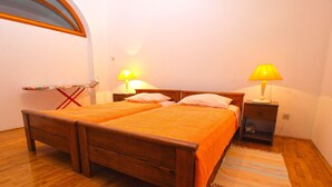 Una cama doble con dos almohadas, suelo de madera, mesita de noche con lámpara y una plancha pequeña.