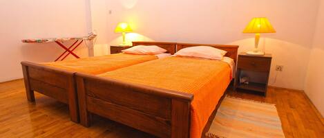 A double bed with two pillows, a wooden floor, a nightstand with a lamp, and a small ironing board.