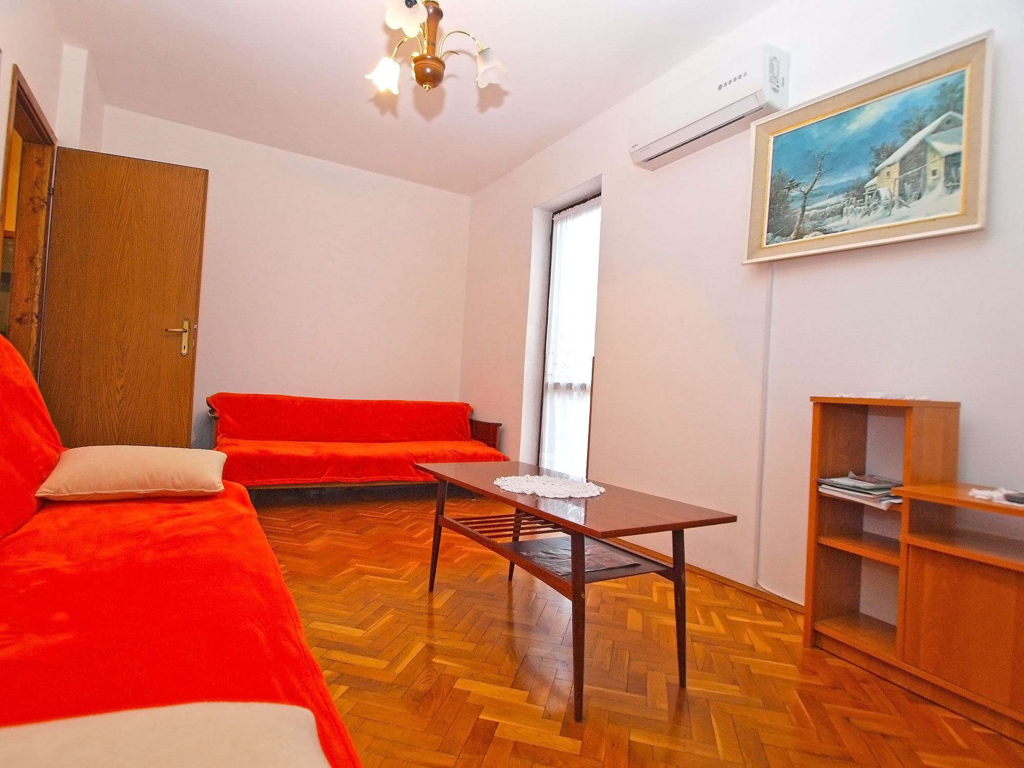 A room with a wooden herringbone floor, a red sofa, a wooden coffee table, a wooden bookshelf, and a painting on the wall.