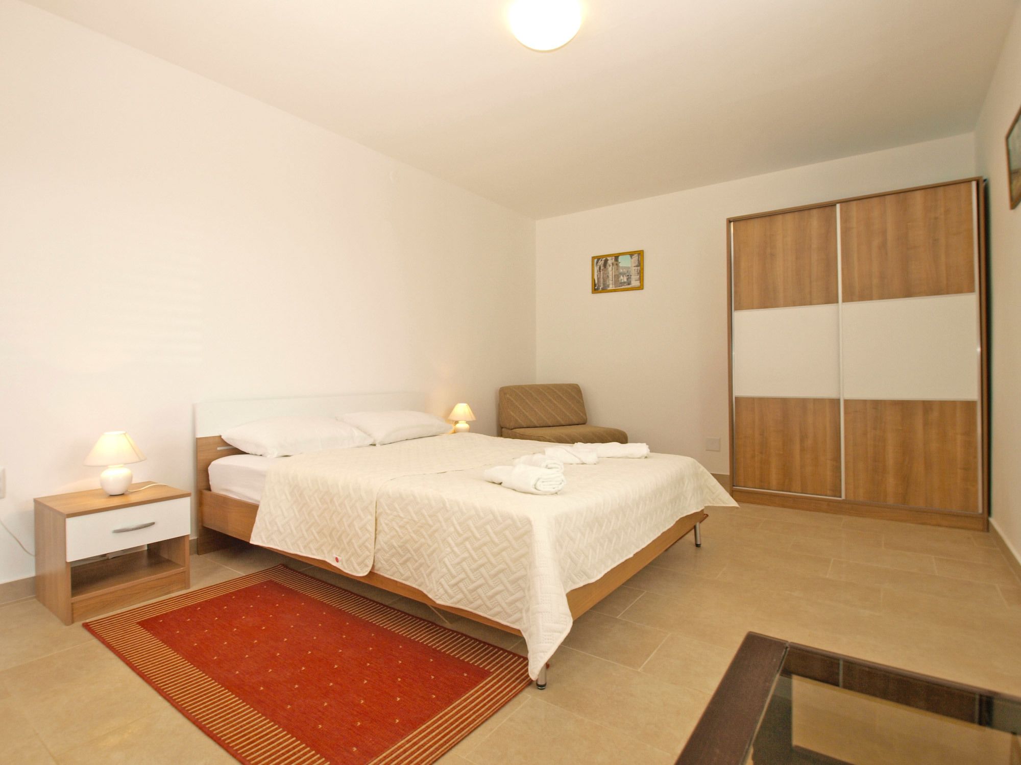 A bedroom with a bed, nightstands, a red rug, a wooden wardrobe, and a painting on the wall.