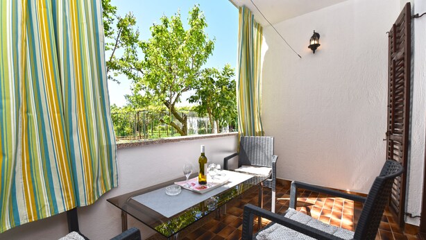 A balcony with a glass table, two chairs, a bottle of wine, and a view of a tree and fence. - Apartment 1230 (Pula)