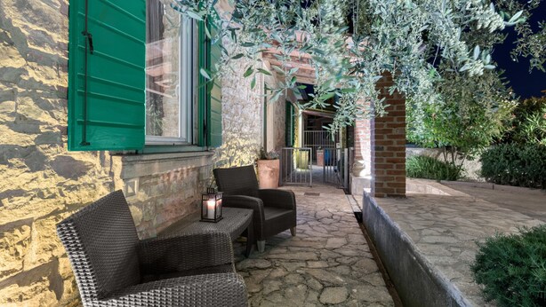 A stone-floored patio with wicker furniture and a lantern, overlooking a garden with olive trees and a brick wall. - House 1231 (Pula)