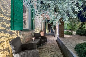 A stone-floored patio with wicker furniture and a lantern, overlooking a garden with olive trees and a brick wall.