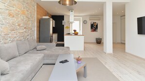 A modern living room with a stone wall, a grey sofa, a white coffee table, and a kitchen area in the background. - House 1231 (Pula)