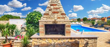 Una chimenea de piedra al aire libre, con una piscina y sillones de descanso al fondo.