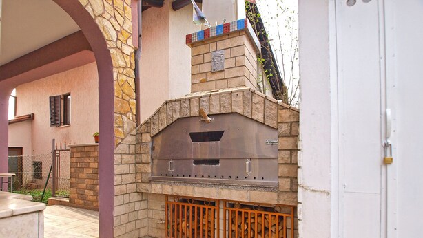 An outdoor barbecue area with a stone and brick fireplace, a tiled patio, and a metal grill enclosure. - Apartment 1265 (Fazana)