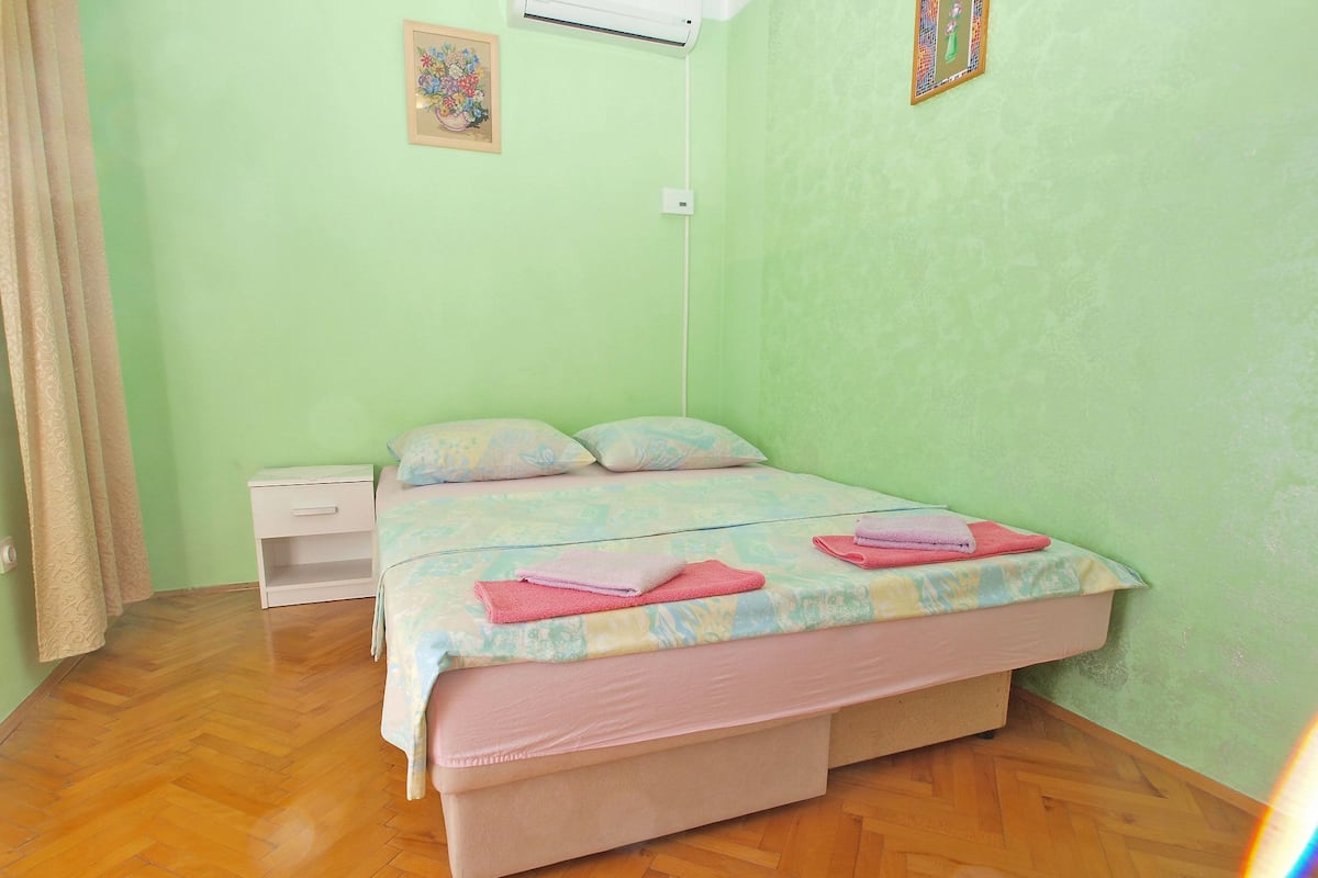 A bedroom with a bed, a nightstand, a wall-mounted air conditioner, and a window with curtains.