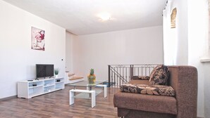 A living room with a brown sofa, a glass coffee table, a TV on a white stand, and a staircase with a metal railing. - House 1277 (Visnjan)