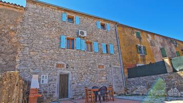 Un edificio de piedra con una puerta y ventanas, un pequeño espacio al aire libre con mesas y sillas, y un camino de grava.