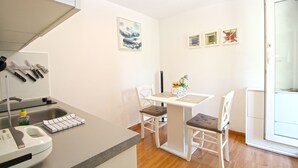 A small kitchenette with a sink, a kettle, and a set of knives. There is a table with two chairs and a vase with flowers. The room has white walls and a wooden floor. - Apartment 1278 (Pula)