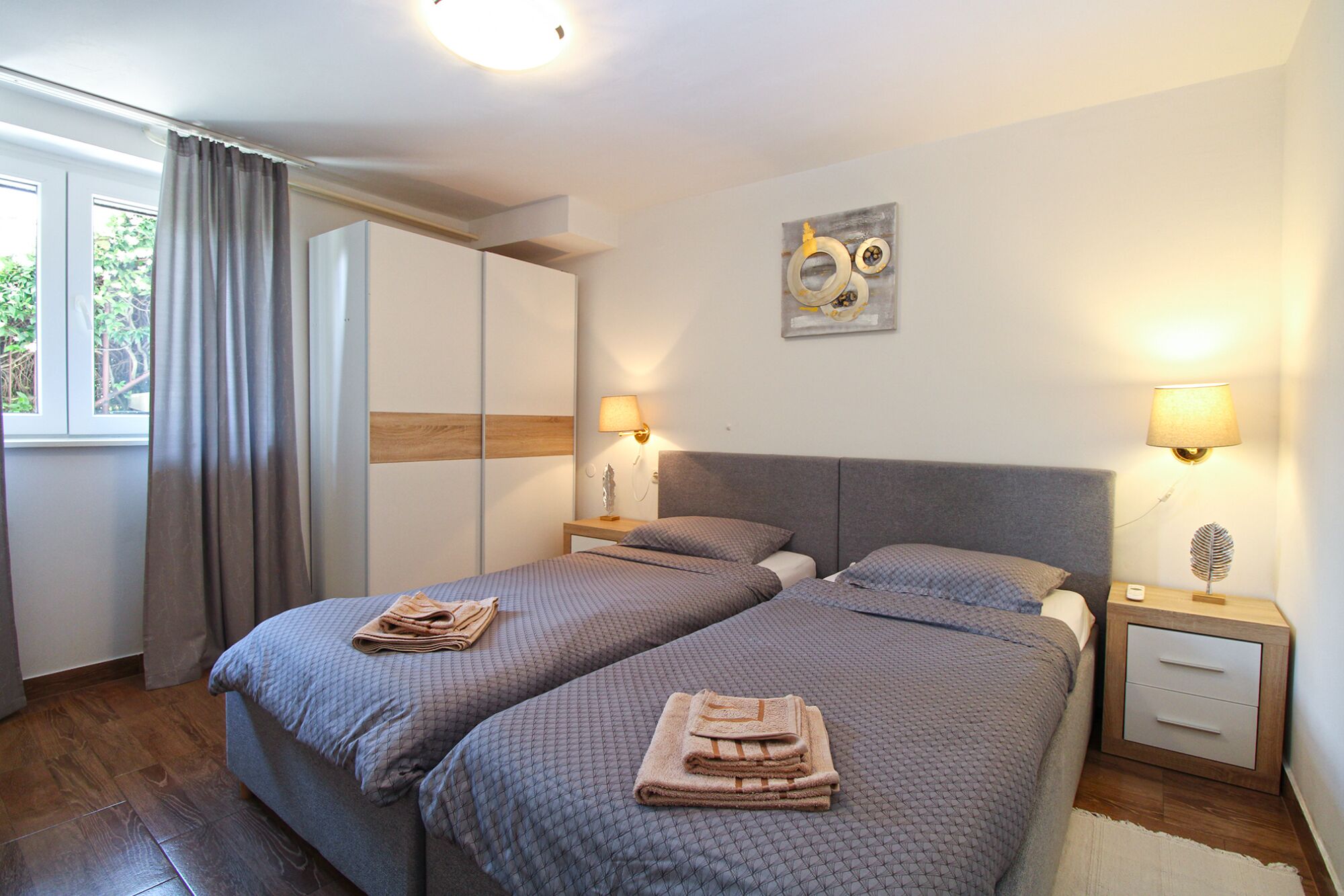 A bedroom with two beds, a wardrobe, a bedside table with a lamp, and a window with curtains.