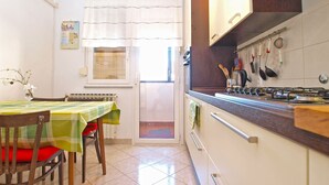 A compact kitchen with a dining area, featuring a table with a green and white checkered tablecloth, wooden chairs, and a window with white curtains. - Apartment 1286 (Pula)