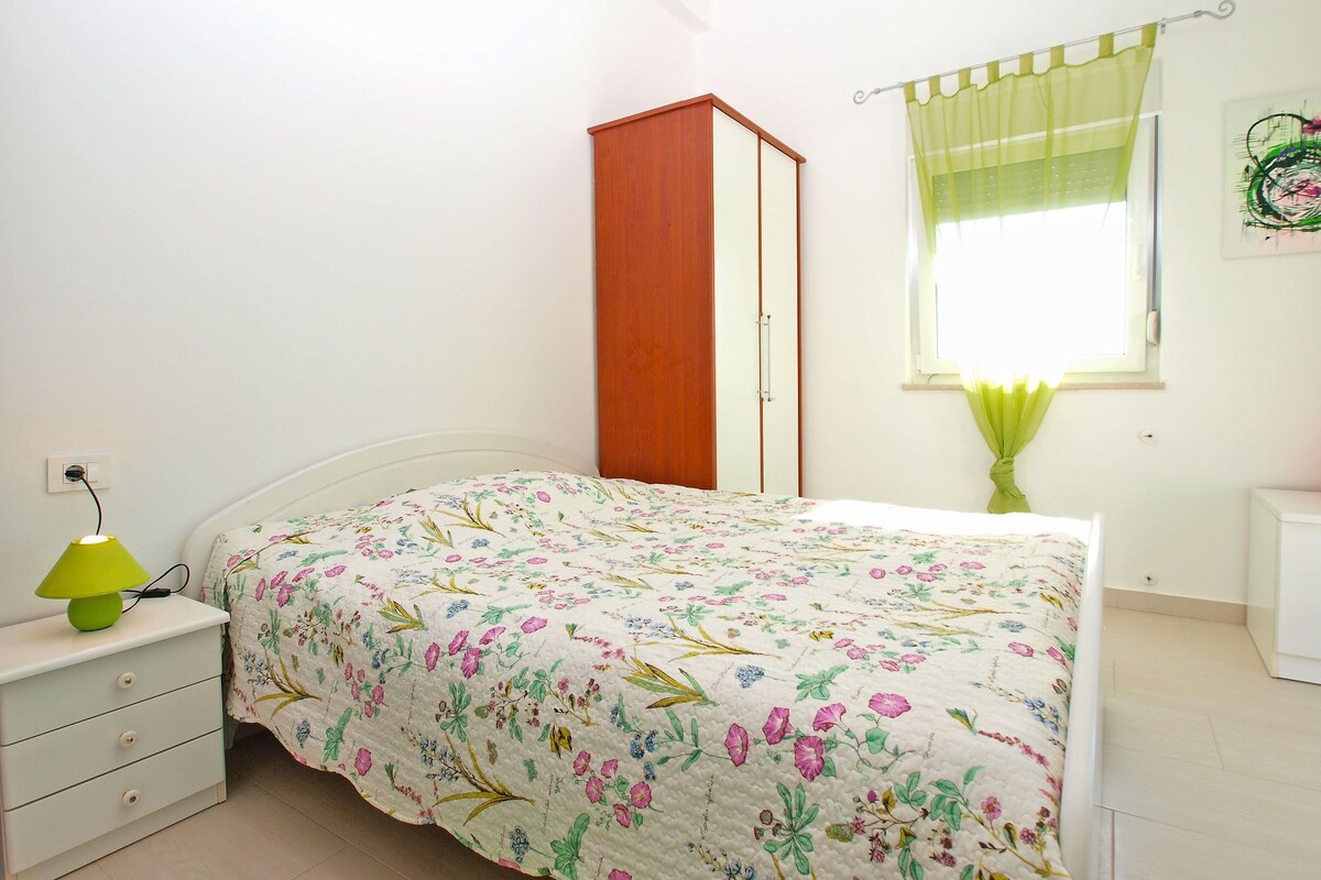 A bedroom with a bed, a nightstand, a wardrobe, a window with curtains, and a wall-mounted light switch.