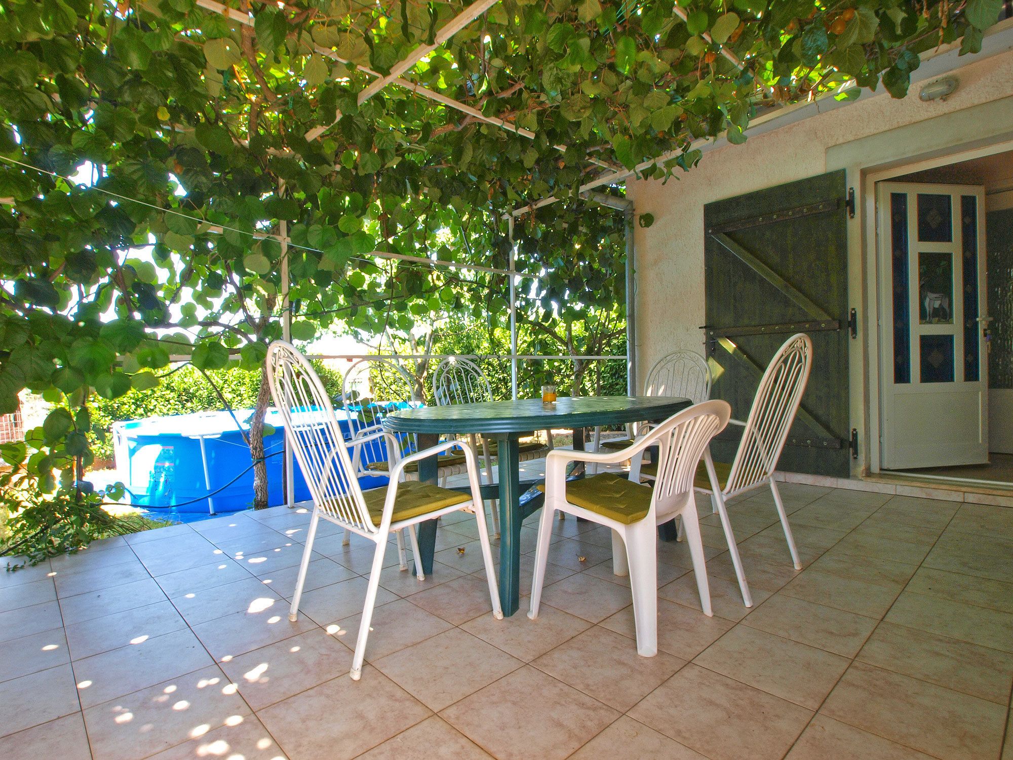 Een terras buiten met een tafel en stoelen, omgeven door groen en een blauwe constructie op de achtergrond.