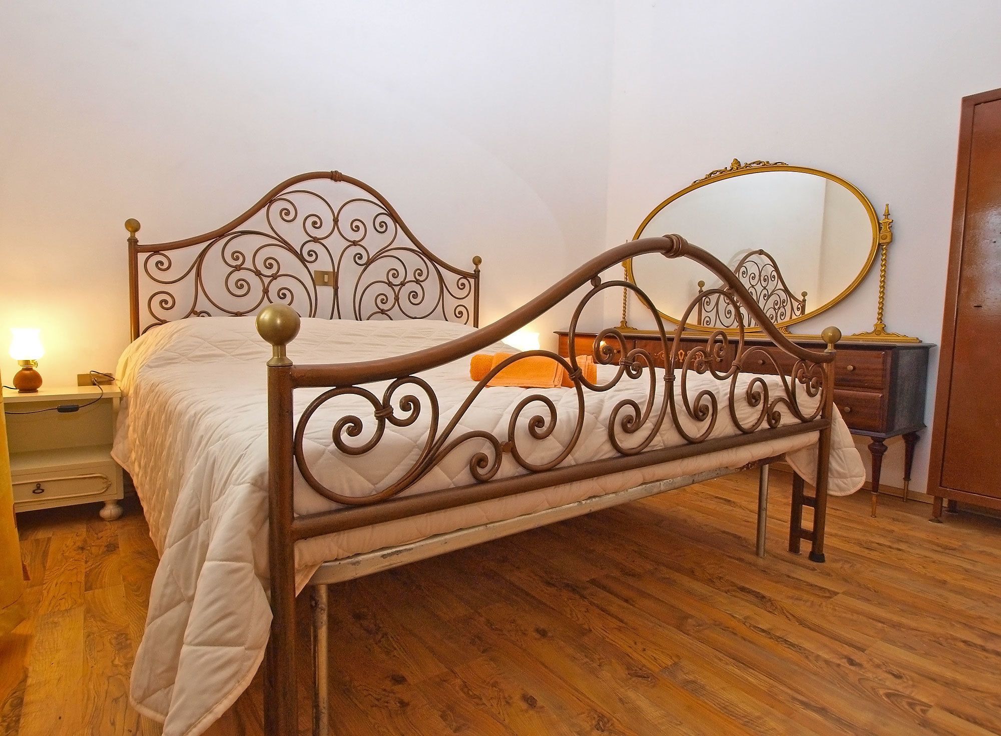 A bed with a metal frame and a headboard, a bedside table with a lamp, and a dresser with a mirror.
