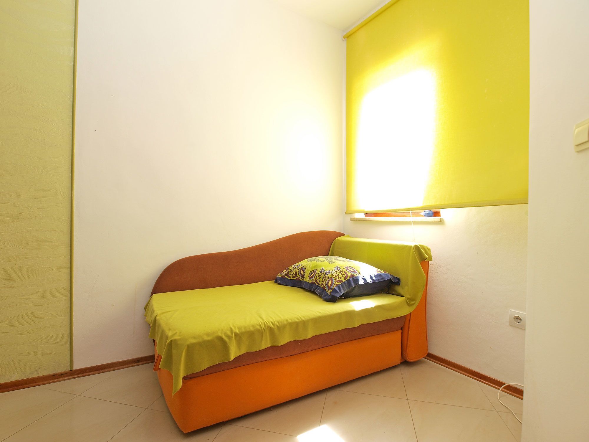 A small, single-bed room with a yellow wall, a window with a yellow blind, and a power outlet on the wall.
