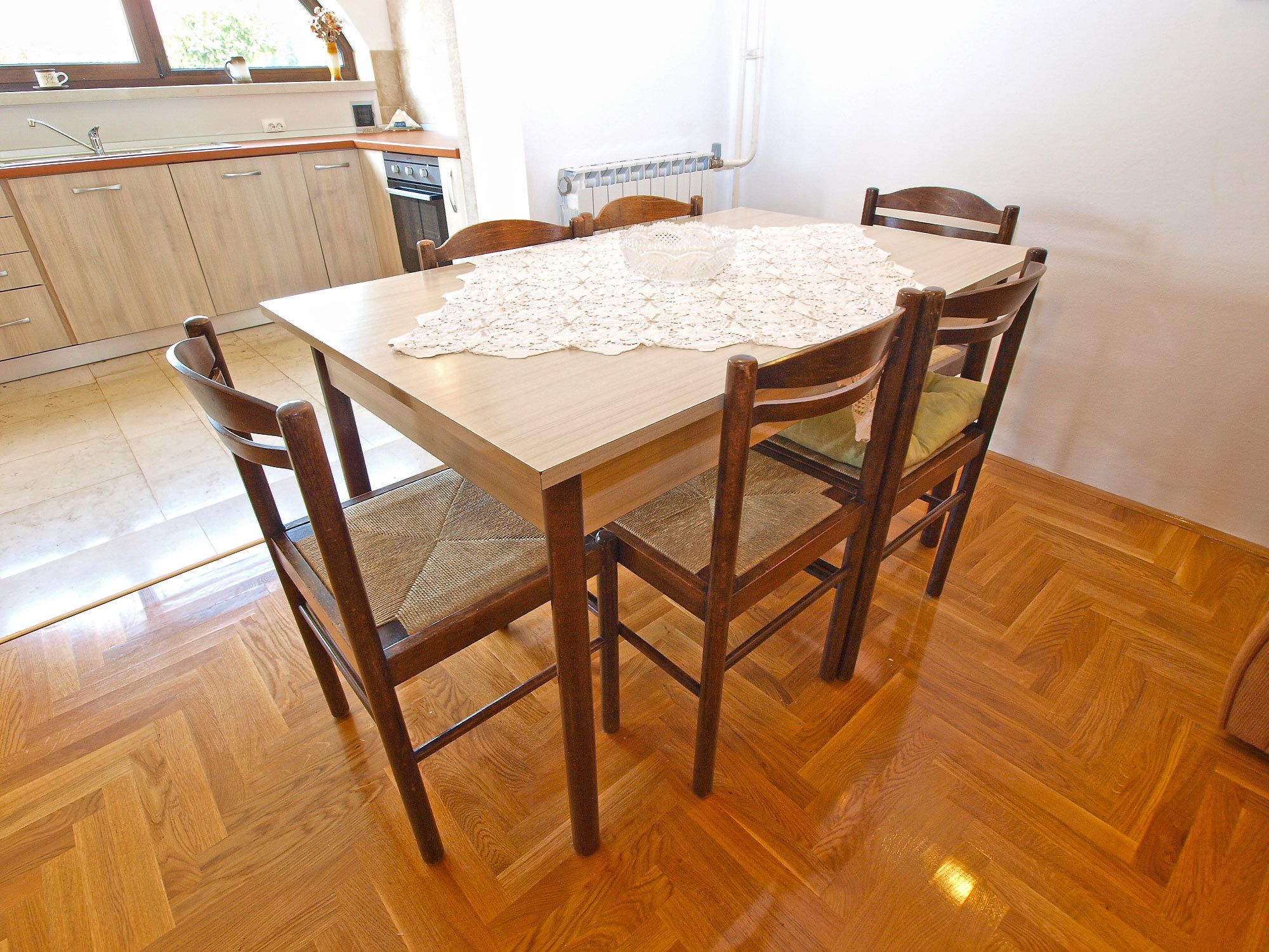 A dining table with a lace tablecloth, wooden chairs, and a kitchen in the background.