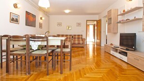 Una sala de estar con piso de madera en patrón de espiga, una mesa de comedor con sillas, un sofá, un televisor y una puerta que da a otra habitación.