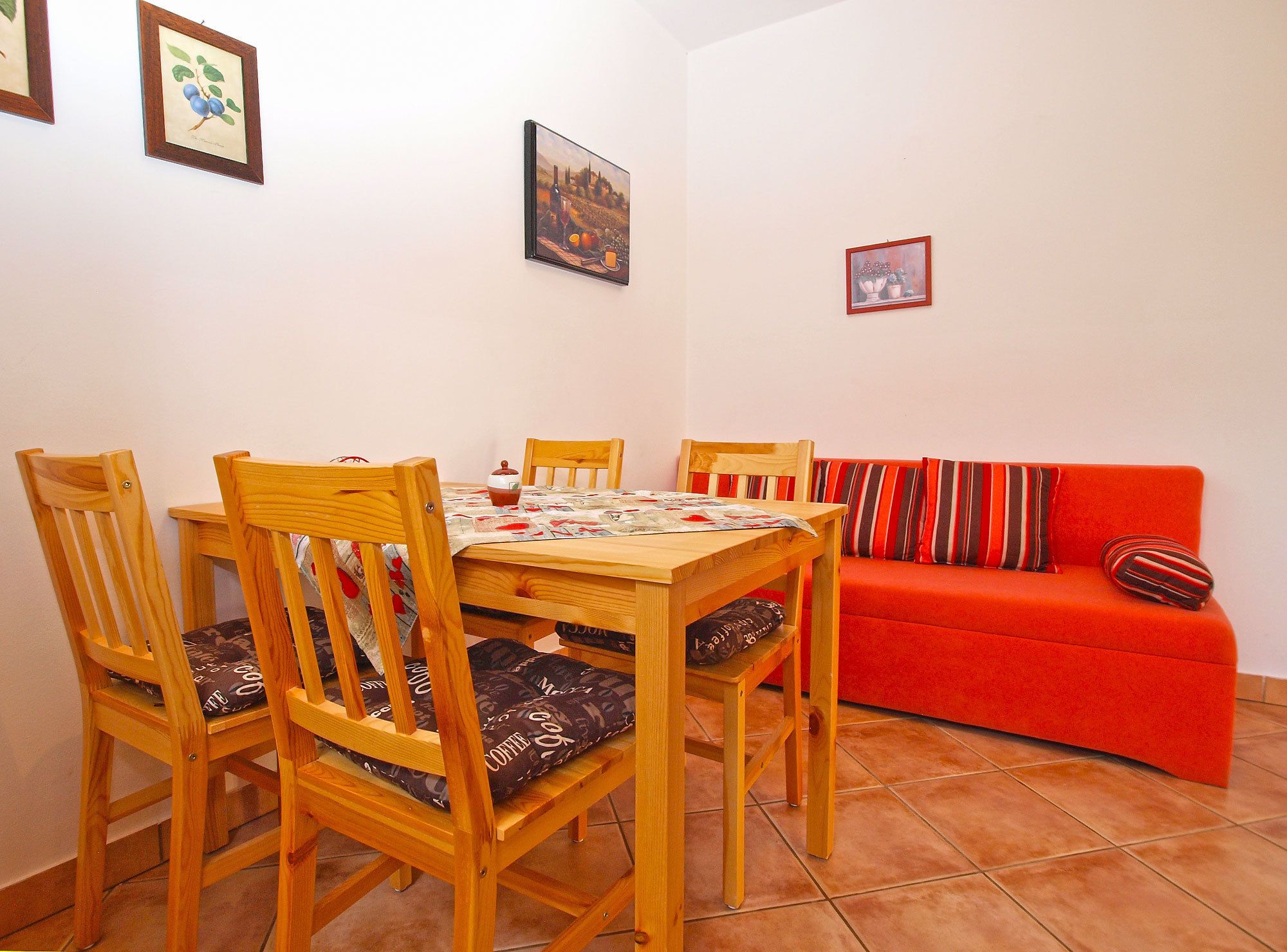 A dining area with a wooden table and chairs, a red sofa with striped cushions, and framed artwork on the walls.
